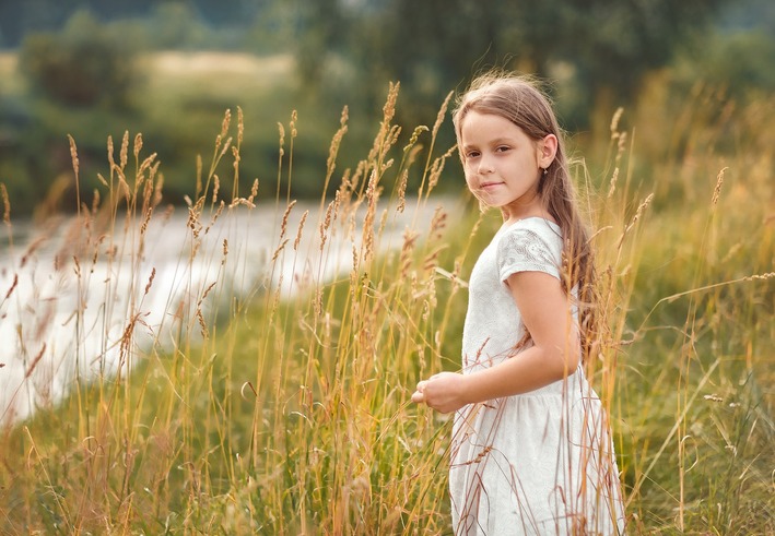 A little girl, Alice, stands in a field next to a lake looking over her shoulder toward the camera