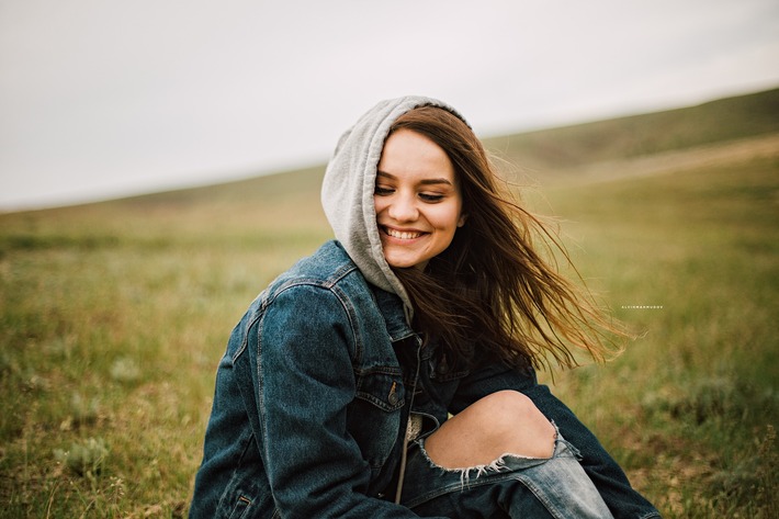 A young woman wearing a grey hoodie, ripped jeans, and denim jacket sits in a grass field, the wind blowing in her long brown hair. Her eyes are cast downward and she is smiling. (Image courtesy of pixabay).