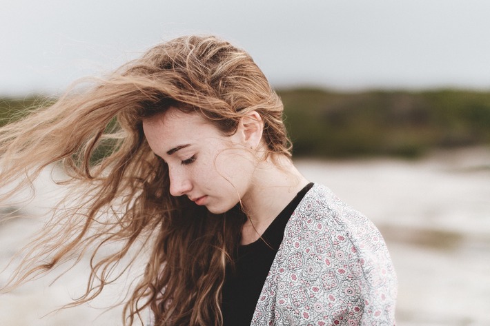 A young woman stands on the beach looking crestfallen, the wind blowing her hair wildly.